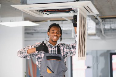 Genç Afro-Amerikan HVK teknisyeni kaset tipi klimayı tamir ediyor ve baş parmaklarını hareket ettiriyor.