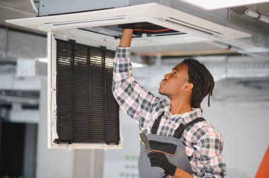 Genç Afrikalı Amerikalı erkek teknisyen havalandırma sistemini tamir ediyor, havalandırma sistemi üzerinde bakım yapıyor.