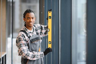 Genç gülümseyen Afrikalı Amerikalı tamirci modern binaya pencereler yerleştirirken balon seviyesini kullanıyor