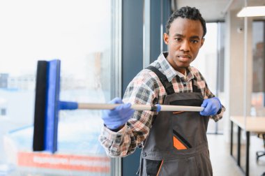 Genç Afro-Amerikan erkek temizlikçi modern bir ofiste cam silici ile camları yıkıyor, bedava parlaklık ve hijyen sağlıyor.