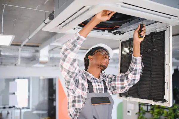 Genç Afro-Amerikan bakım işçisi bir tornavidayla havalandırma sistemini tamir ediyor. İçerideki en uygun soğutma ve hava akışını sağlıyor.