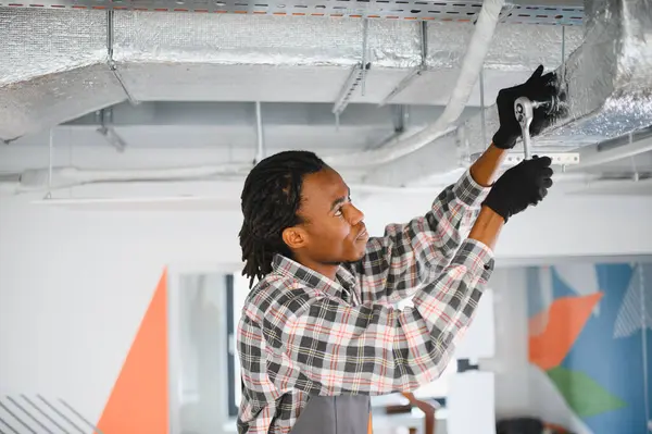 Eldiven ve önlük giyen genç bir Afro-Amerikan işçi. İngiliz anahtarı kullanıyor. Modern bir ofiste havalandırma borusunu tamir ediyor.