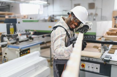 Gülümseyen Afro-Amerikan marangoz ahşap bir kalası incelerken mobilya fabrikasında çalışıyor, zanaat ve beceri sergiliyor.