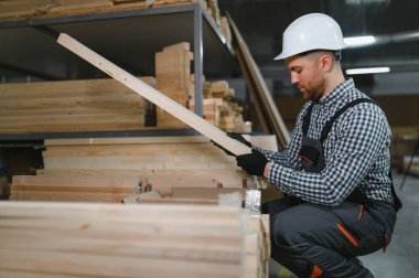 Marangoz bir güvenlik kaskı takıyor ve hareketli bir mobilya fabrikasındaki ahşap kalasları titizlikle inceliyor.
