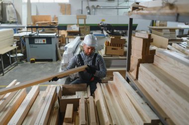 Marangoz güvenlik kaskı takıyor. Tahta ve makinelerle çevrili bir mobilya fabrikasında ahşap kalasları kontrol ediyor.