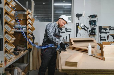 Marangoz, aletler ve malzemelerle çevrili bir mobilya fabrikasında pnömatik bir aletle tahta işlemeye konsantre oluyor.