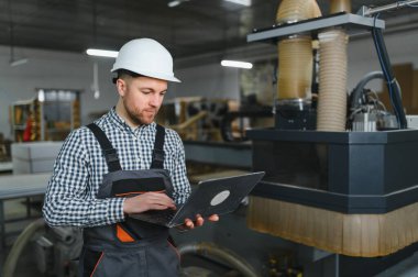 Marangoz güvenlik kaskı takıyor ve dizüstü bilgisayar kullanıyor. Mobilya fabrikasında bir CNC makinesini yönetiyor.