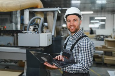 Marangoz, mobilya fabrikasındaki bir Cnc makinesini kontrol etmek için dizüstü bilgisayar kullanıyor, üretim sürecini optimize ediyor ve kaliteyi sağlıyor.