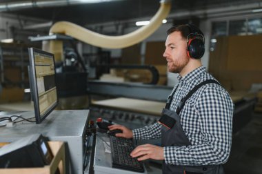 Marangoz kulaklık takıyor ve hareketli bir mobilya fabrikasında ahşap yönlendirici kullanıyor. Yetenekli işçilik ve modern teknoloji sergiliyor.