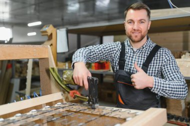 Marangoz, pnömatik zımba tabancası kullanarak ahşap kanepe çerçevesi monte ediyor ve mobilya fabrikasında baş parmak hareketlerini gösteriyor.