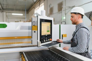 Bir mobilya fabrikasında güvenlik kaskı takan marangoz, ahşap işleme yazılımı programlıyor.