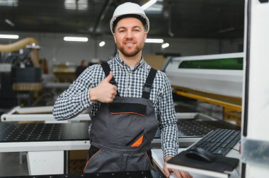 Mobilya fabrikasında otomatikleştirilmiş ahşap işçiliği makinesinin yanında dururken gülümseyen marangozun baş parmağını gösteren portresi