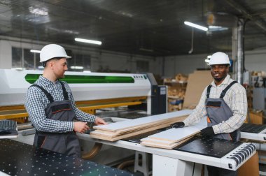 İki çoklu etnik marangoz bir mobilya fabrikasında ahşap tahtalarla çalışıyor, hassas kesim ve montaj için bir cnc makinesi kullanıyorlar.