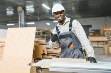 Kendine güvenen Afrikalı Amerikalı marangoz, mobilya fabrikasındaki modern makineler üzerine ahşap panelle çalışırken baş parmak hareketi yapıyor.