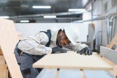 Marangoz koruyucu iş giysisi ve şapka takıyor ahşap kalası dikkatlice inceliyor, mobilya üretiminde kalite kontrolü sağlıyor