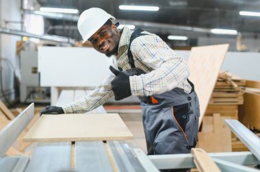 Gülümseyen marangoz, mobilya fabrikasında ahşap paneli makinelerle keserken baş parmağını kaldırıyor.