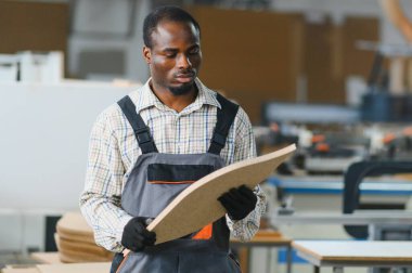 Tahta bir paneli inceleyen bir mobilya fabrikasında çalışan Afrikalı Amerikalı işçi. Arka planda cnc makineleri olan sanayi ayarı
