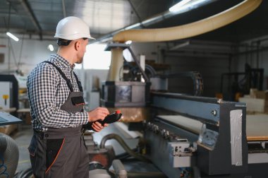 Marangoz güvenlik kaskı ve tulum giyer, bir mobilya fabrikasında CNC makinesini çalıştırır, süreci uzaktan kumandayla kontrol eder.