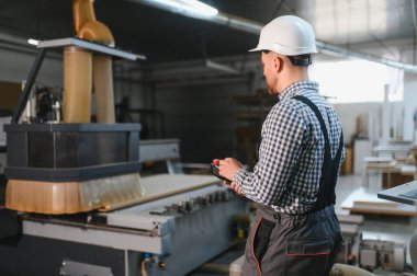 Marangoz, güvenlik kaskı ve tulumu takıyor. Mobilya fabrikasında ahşap yönlendirici kullanıyor. Elinde uzaktan kumanda var.