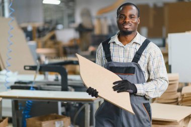 Gülümseyen Afro-Amerikan marangoz elinde tahta bir kalasla kalabalık bir mobilya fabrikasında çalışırken, zanaat ve beceri gösterisi yapıyor.