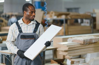 Marangoz bir mobilya fabrikasında ahşap bir kalas inceliyor, montajdan önce kalite kontrolüne odaklanıyor.