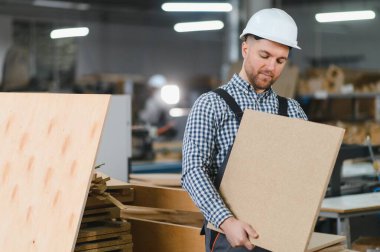 Marangoz kalın bir şapka takıyor ve tahta bir tahta tutuyor. Yoğun bir fabrika ortamında mobilya üretiyor.
