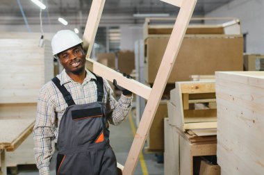 Meşgul bir mobilya fabrikasında çalışırken ahşap bir çerçeve taşıyan gülümseyen marangoz, uzmanlık ve beceri gösterisi yapıyor.