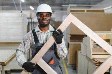 Gülümseyen marangoz, mobilya fabrikasında çalışırken ahşap bir çerçeve tutuyor. Mobilya üretiminde beceri ve zanaat sergiliyor.