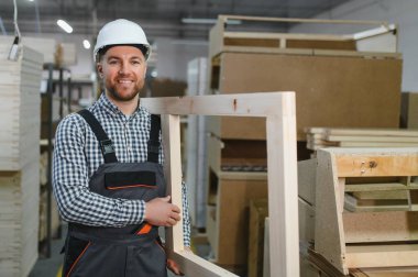 Meşgul bir mobilya fabrikasında çalışırken ahşap bir çerçeve tutan gülümseyen marangoz, marangozluk ve adanmışlık sergiliyor.