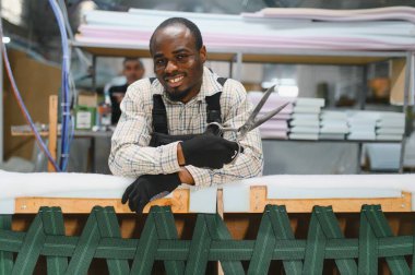 Bir mobilya fabrikasında kanepede çalışırken elinde makasla gülümseyen Afrikalı bir Amerikalı zanaatkarın portresi.