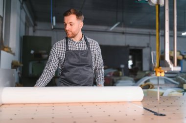 Marangoz tezgahın üzerine büyük beyaz kumaş rulo açıyor, mobilya fabrikasında otomatik kesim makinesi için malzeme hazırlıyor.