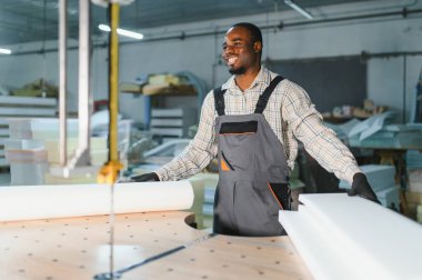 Gülümseyen Afro-Amerikan işçiler mobilya fabrikasındaki testereyi kullanarak kanepe üretimi için köpük kesiyorlar.