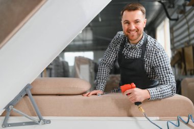 Marangoz bir mobilya fabrikasında pnömatik tornavida kullanarak bir kanepe kuruyor, kameraya doğru gülümsüyor.