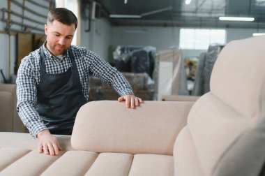 Marangoz, bir mobilya fabrikasında yeni yapılmış bir kanepenin döşemelerini inceliyor. Üretimde kalite ve detaya dikkat çekiyor.