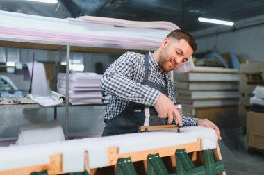 Yetenekli marangoz, ahşap bir kanepe çerçevesine kumaş bağlamak için pnömatik zımba tabancası kullanır ve yoğun bir mobilya fabrikasında özenle çalışır.
