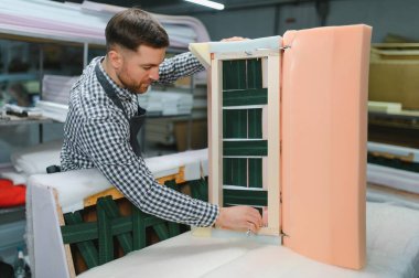 Marangoz mobilya fabrikasında çalışıyor. Kanepenin ahşap çerçevesini elastik ağ ve köpük dolgu malzemesi kullanarak birleştiriyor.