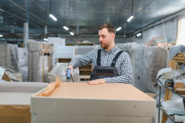 Bir mobilya fabrikasında çalışan marangoz, çivi tabancasıyla bir kanepenin ahşap çerçevesini birleştiriyor. Etrafı malzeme ve bitmiş ürünlerle çevrili.