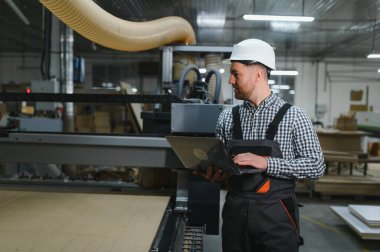 Marangoz dizüstü bilgisayarda çalışıyor, çalışan bir mobilya fabrikasında cnc ahşap kesme makinesini programlıyor, teknolojiyi işçilikle harmanlıyor.