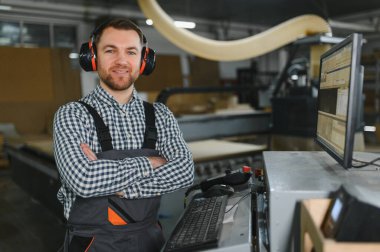 Marangoz kulaklık takıyor ve bir mobilya fabrikasında kollarını kavuşturup gülümsüyor.