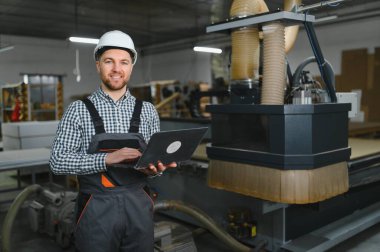 Marangoz sabit şapka takıyor, bir mobilya fabrikasında çalışırken dizüstü bilgisayarı programlıyor ve hassasiyet ve verimliliğe odaklanıyor.