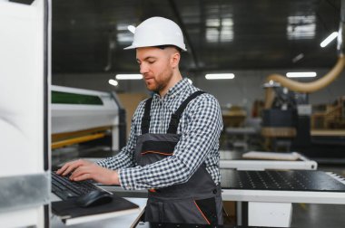 Marangoz, modern bir mobilya fabrikasında çalışırken güvenlik kaskı takarak CNC makinesini programlıyor ve üretim verimliliğini arttırıyor.