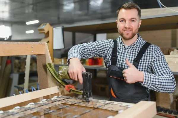 Marangoz, pnömatik zımba tabancası kullanarak ahşap kanepe çerçevesi monte ediyor ve mobilya fabrikasında baş parmak hareketlerini gösteriyor.