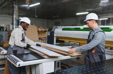 İki marangoz, mobilya fabrikasında çalışan bir makinenin yanında işlenmiş ahşap tahtaları taşırken iş birliği yapıyor.