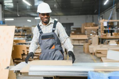 Gülümseyen Afro-Amerikan marangoz mobilya fabrikasındaki modern makine üzerinde ahşap panelle çalışıyor.