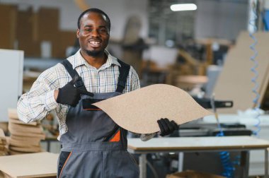 Gülümseyen Afro-Amerikan marangoz, faaliyet ve işçilikle dolu işlek bir mobilya fabrikasında çalışırken bir odun parçasını işaret ediyor.