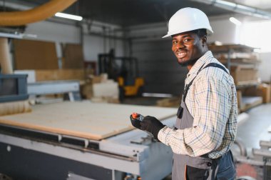 Gülümseyen Afro-Amerikan işçiler mobilya fabrikasında uzaktan kumandayla ahşap bir cnc makinesini kontrol ediyorlar.
