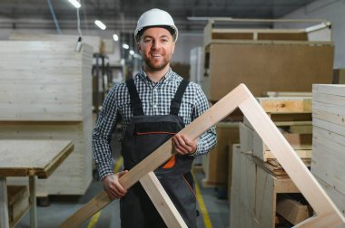 Gülümseyen marangoz ahşap bir çerçeve tutuyor, çalışkan bir mobilya fabrikasında özenle çalışıyor, marangozluk ve adanmışlık sergiliyor.