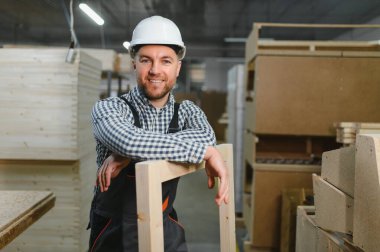 Marangoz, mobilya fabrikasında kasketli ve tulumlu, gülümseyen ve ahşap çerçeveye yaslanmış.