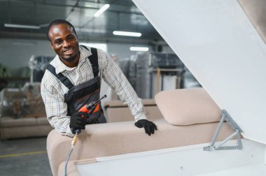 Marangoz, yoğun bir mobilya fabrikasında pnömatik bir tornavidayla kanepe monte ediyor. Yetenekli işçilik ve endüstriyel üretim sergiliyor.