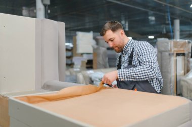 Marangoz bir mobilya fabrikasında yeni mobilyalar üzerine ambalaj kağıdını dikkatlice kesiyor, hassasiyet ve kaliteye odaklanıyor.